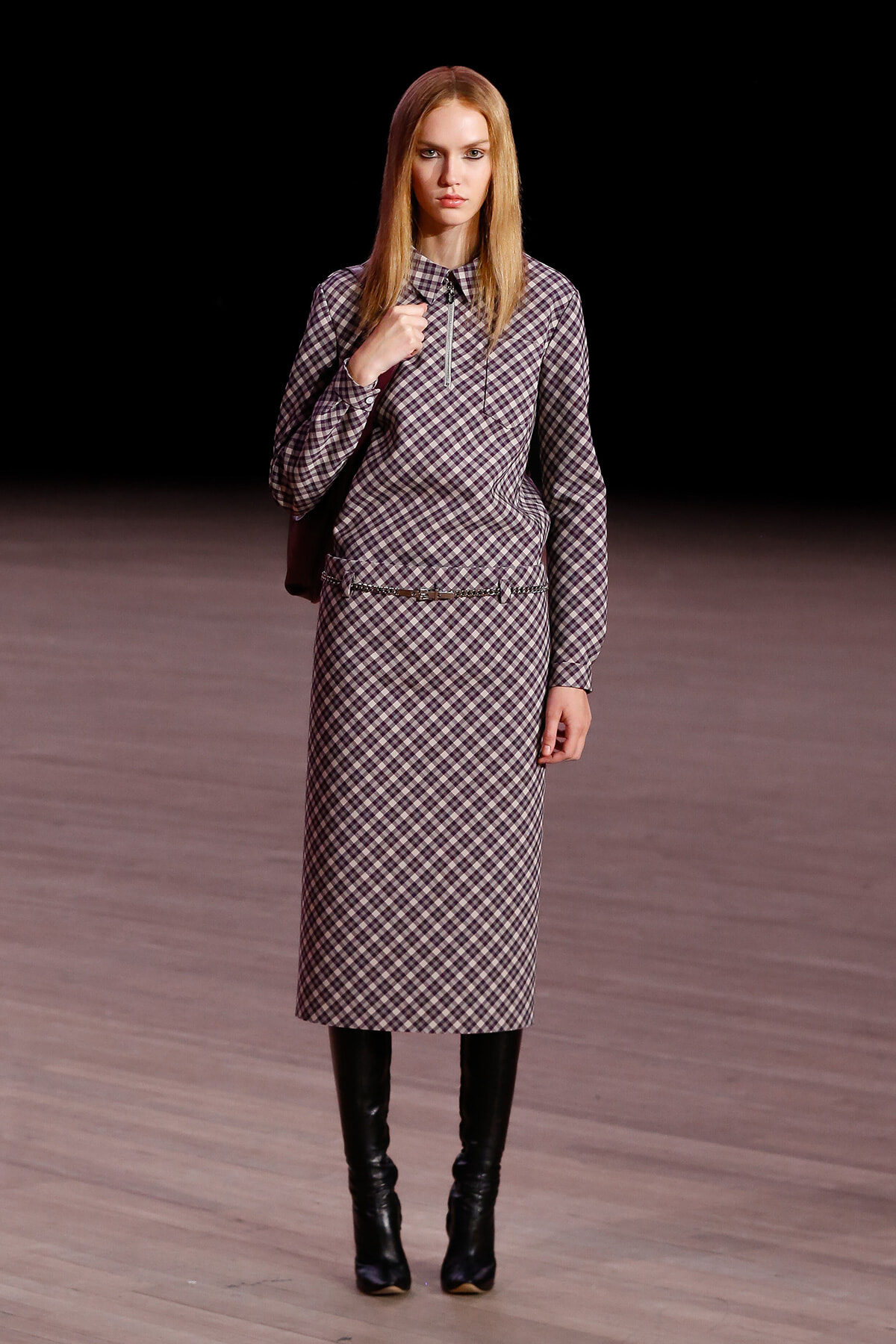 Model on a runway wearing a matching plaid midi dress with long sleeves, a zip collar, and a slim belt, paired with knee-high black boots against a dark background.