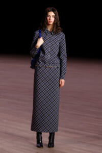 Female model on a runway wearing a matching blue plaid zip-up jacket and ankle-length skirt, with a blue bag over her shoulder.