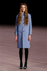 Model on a runway wearing a light blue knee-length trench coat, black tights, and black-and-white heeled shoes; dark backdrop and wooden floor.
