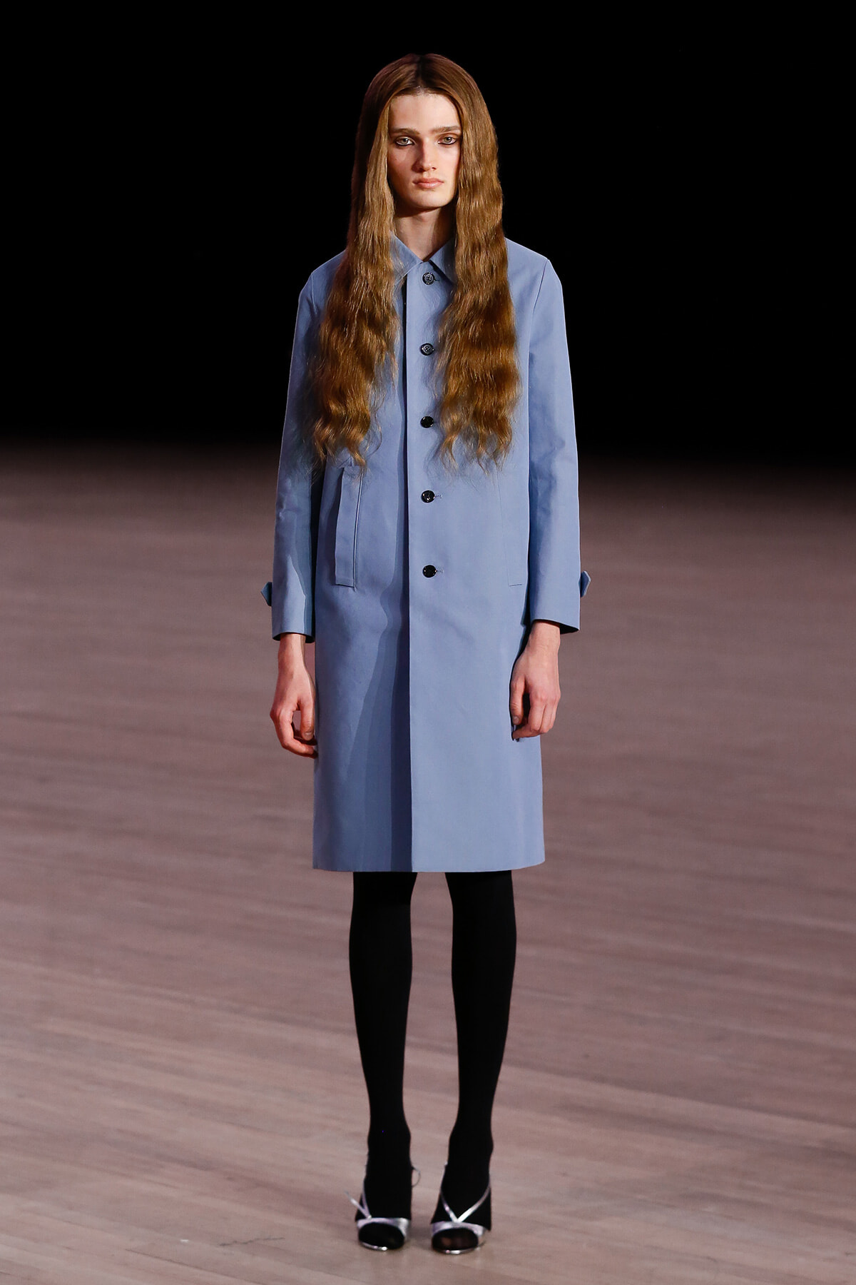 Model on a runway wearing a light blue knee-length trench coat, black tights, and black-and-white heeled shoes; dark backdrop and wooden floor.