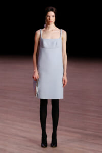Runway model in a pale blue square-neck dress with thin straps, black tights, and a metallic silver clutch.