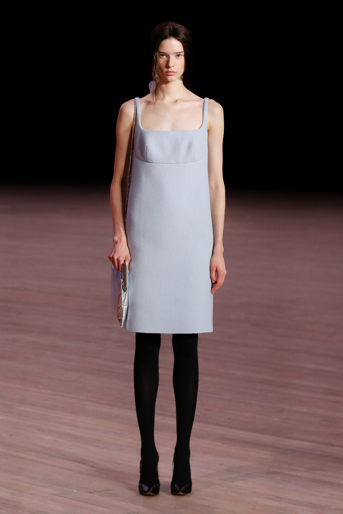 Runway model in a pale blue square-neck dress with thin straps, black tights, and a metallic silver clutch.