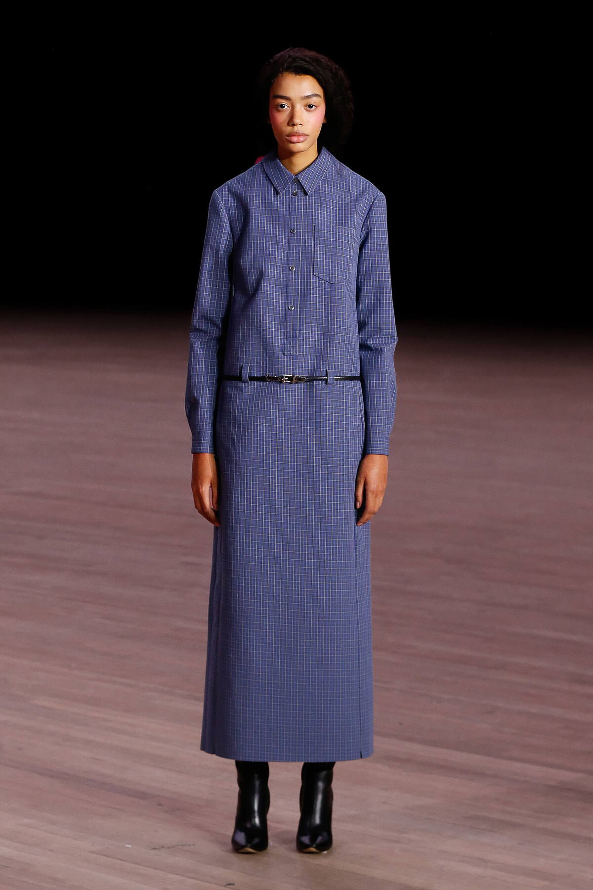 Full-length model in a blue plaid shirt dress with a thin belt and black ankle boots on a runway stage.