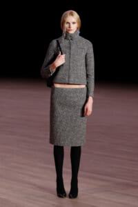 Model on a runway wearing a sparkly gray cropped tweed jacket and matching knee-length skirt, black tights and heels, carrying a black shoulder bag.