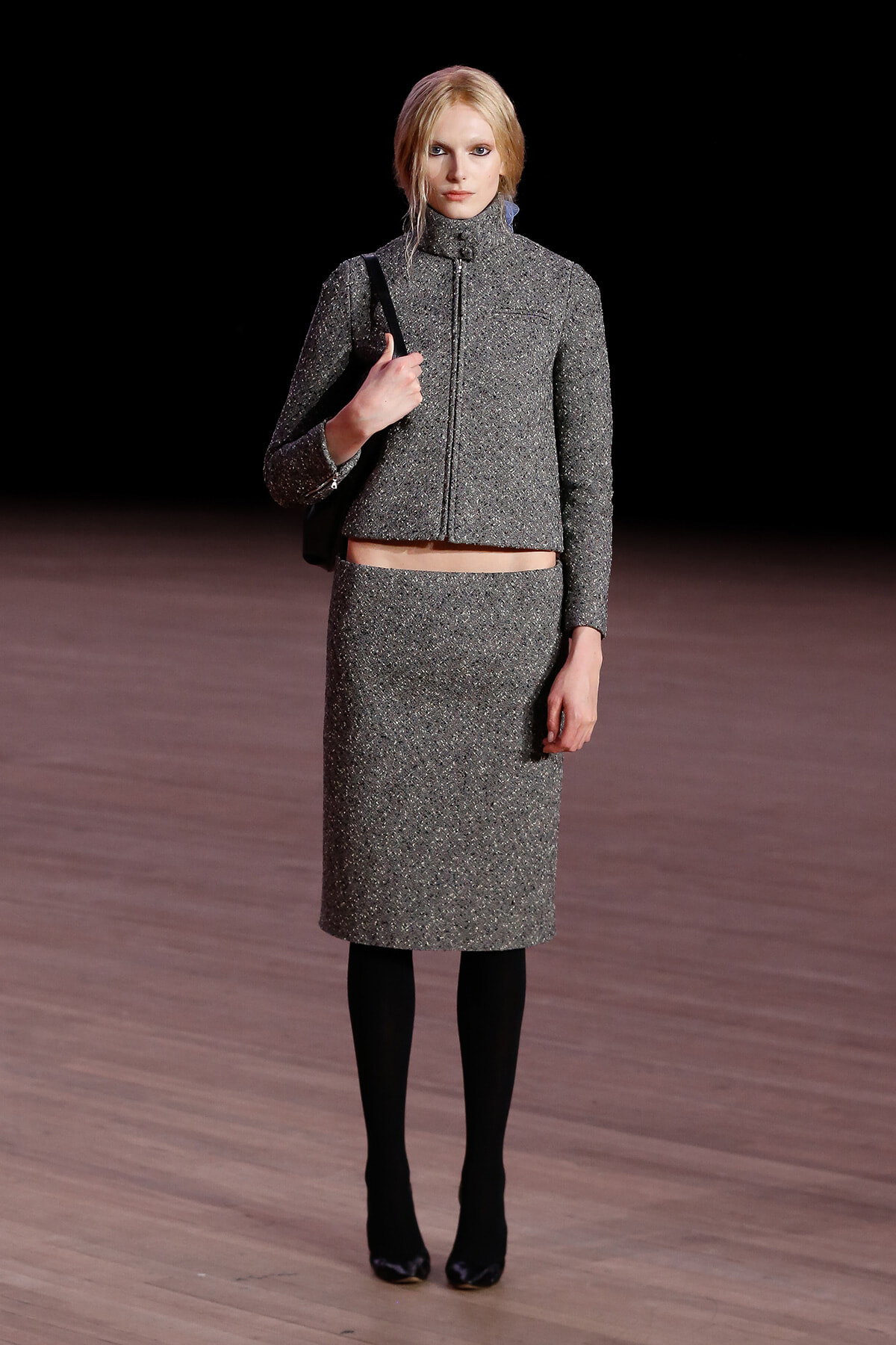 Model on a runway wearing a sparkly gray cropped tweed jacket and matching knee-length skirt, black tights and heels, carrying a black shoulder bag.