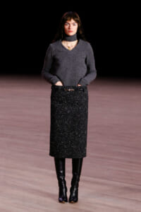 Model on runway wearing a charcoal gray knit sweater with a belted, glittery black midi skirt and tall black boots.
