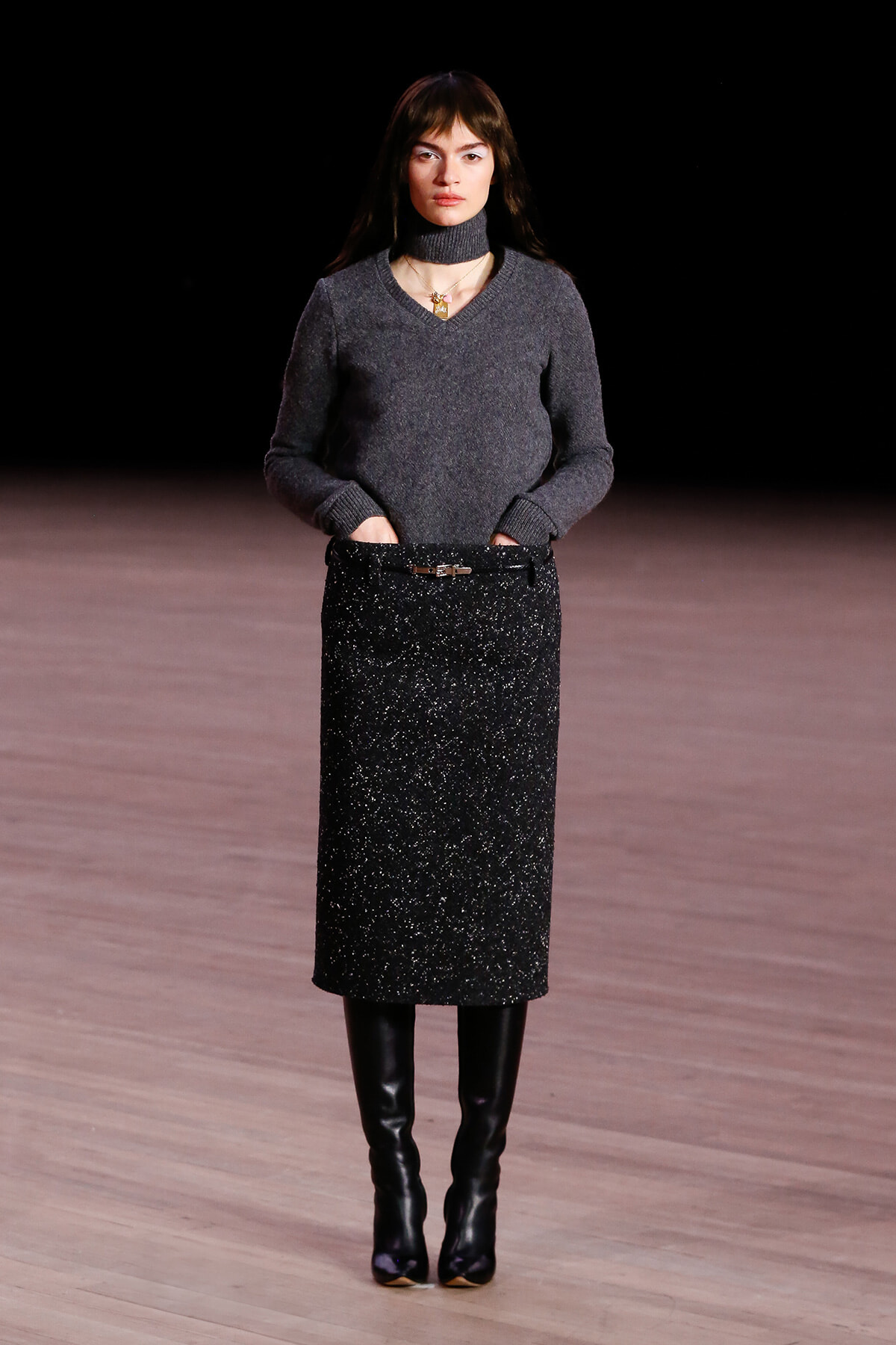 Model on runway wearing a charcoal gray knit sweater with a belted, glittery black midi skirt and tall black boots.