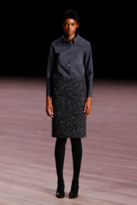 Model standing on a runway in a navy patterned button-down shirt and a black glittery knee-length skirt, with black tights and heels.