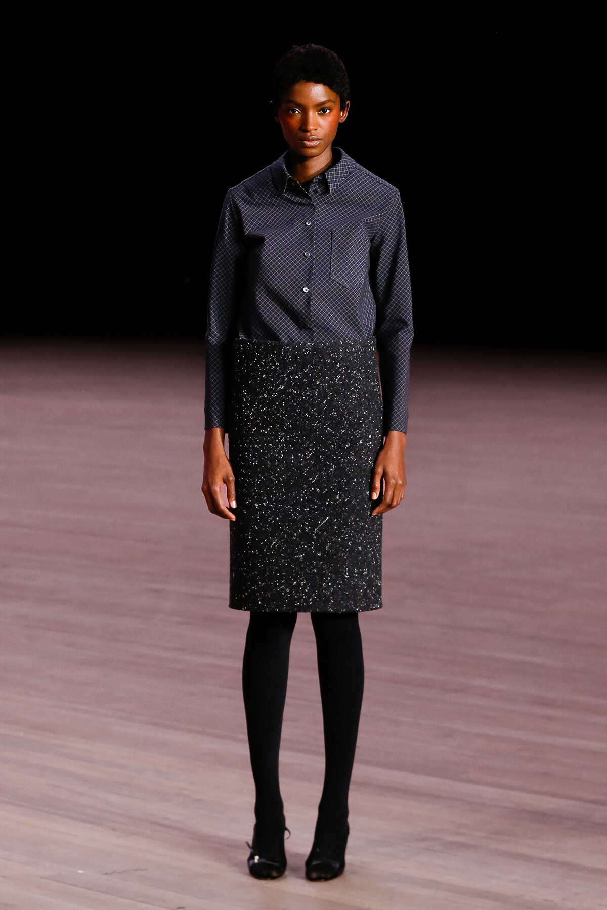 Model standing on a runway in a navy patterned button-down shirt and a black glittery knee-length skirt, with black tights and heels.
