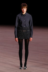 Model walking a runway in a dark checkered button-up shirt tucked into a glittery black mini skirt with tights and white-heeled sandals.