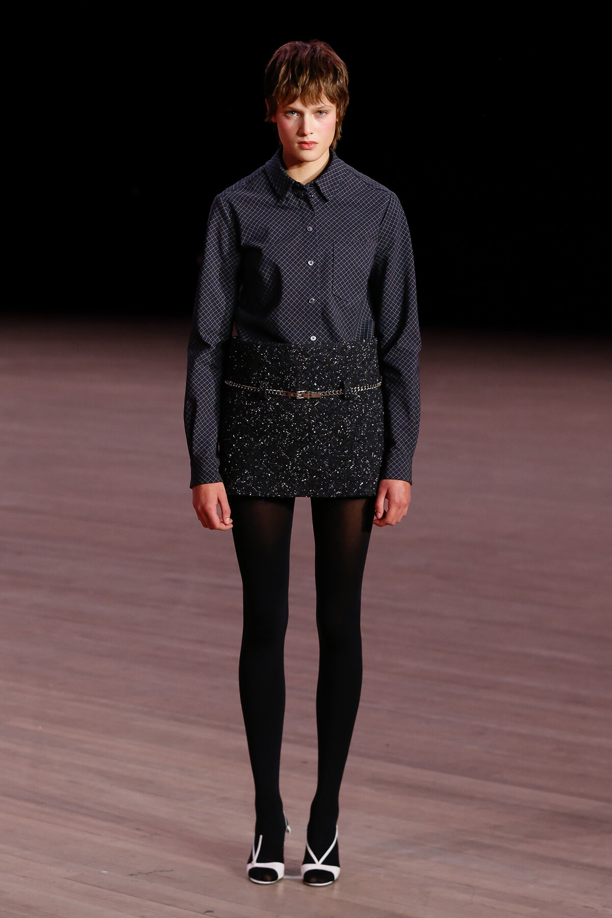 Model walking a runway in a dark checkered button-up shirt tucked into a glittery black mini skirt with tights and white-heeled sandals.