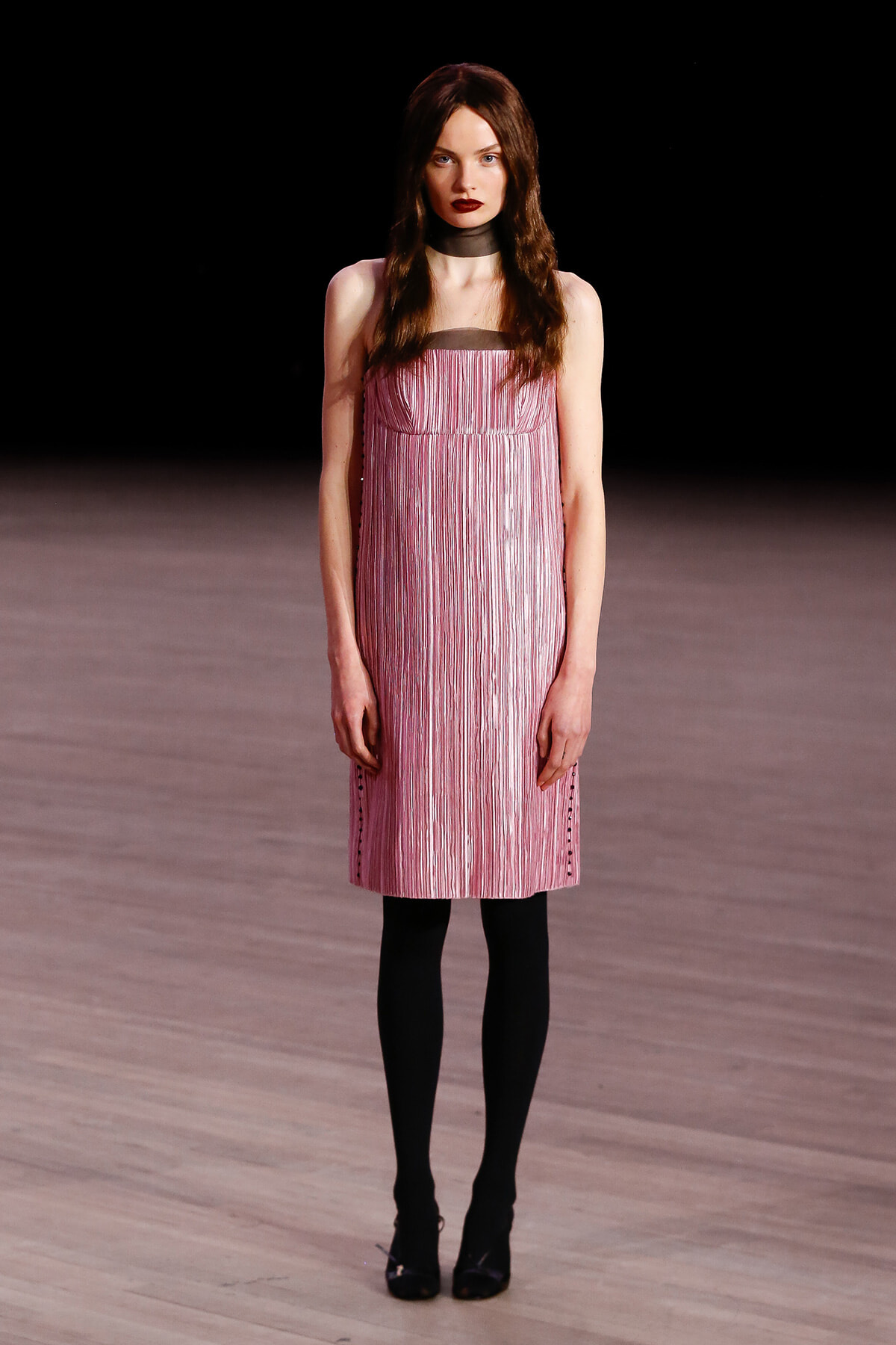 Model standing on a runway in a pink pleated dress with a sheer neckline, black tights and black heels against a dark background.