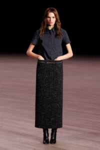Model walking a runway in a dark short-sleeve plaid shirt and a long glittery black skirt with ankle boots.