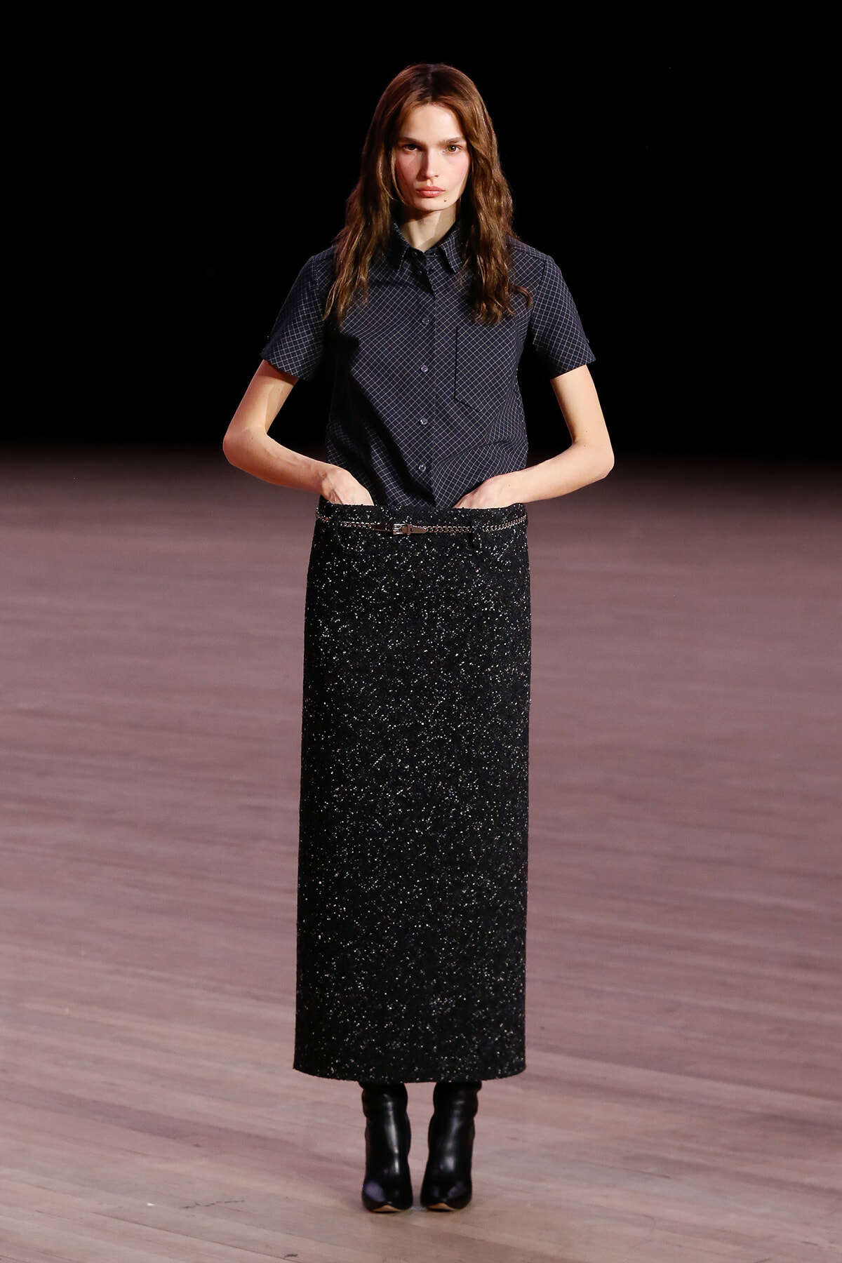 Model walking a runway in a dark short-sleeve plaid shirt and a long glittery black skirt with ankle boots.