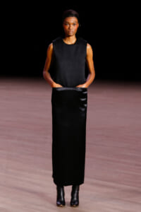 Model wearing a sleeveless black top and long black satin skirt on a runway, hands in pockets, neutral expression.
