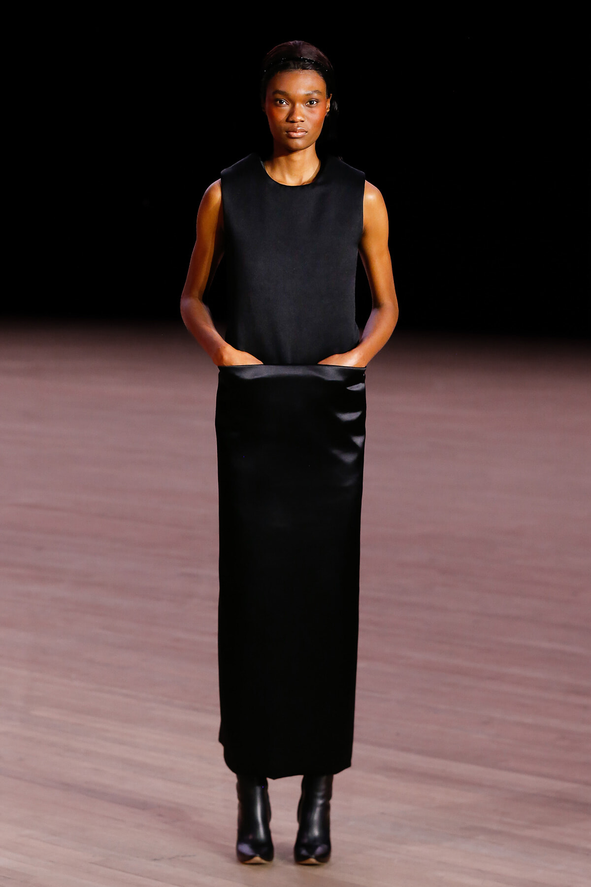 Model wearing a sleeveless black top and long black satin skirt on a runway, hands in pockets, neutral expression.