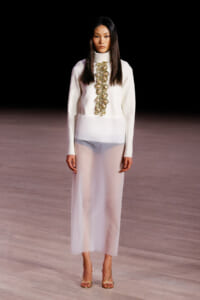Model on a runway wearing a white high-neck top with a gold-embellished vertical appliqué and matching white skirt, gold strappy heels.