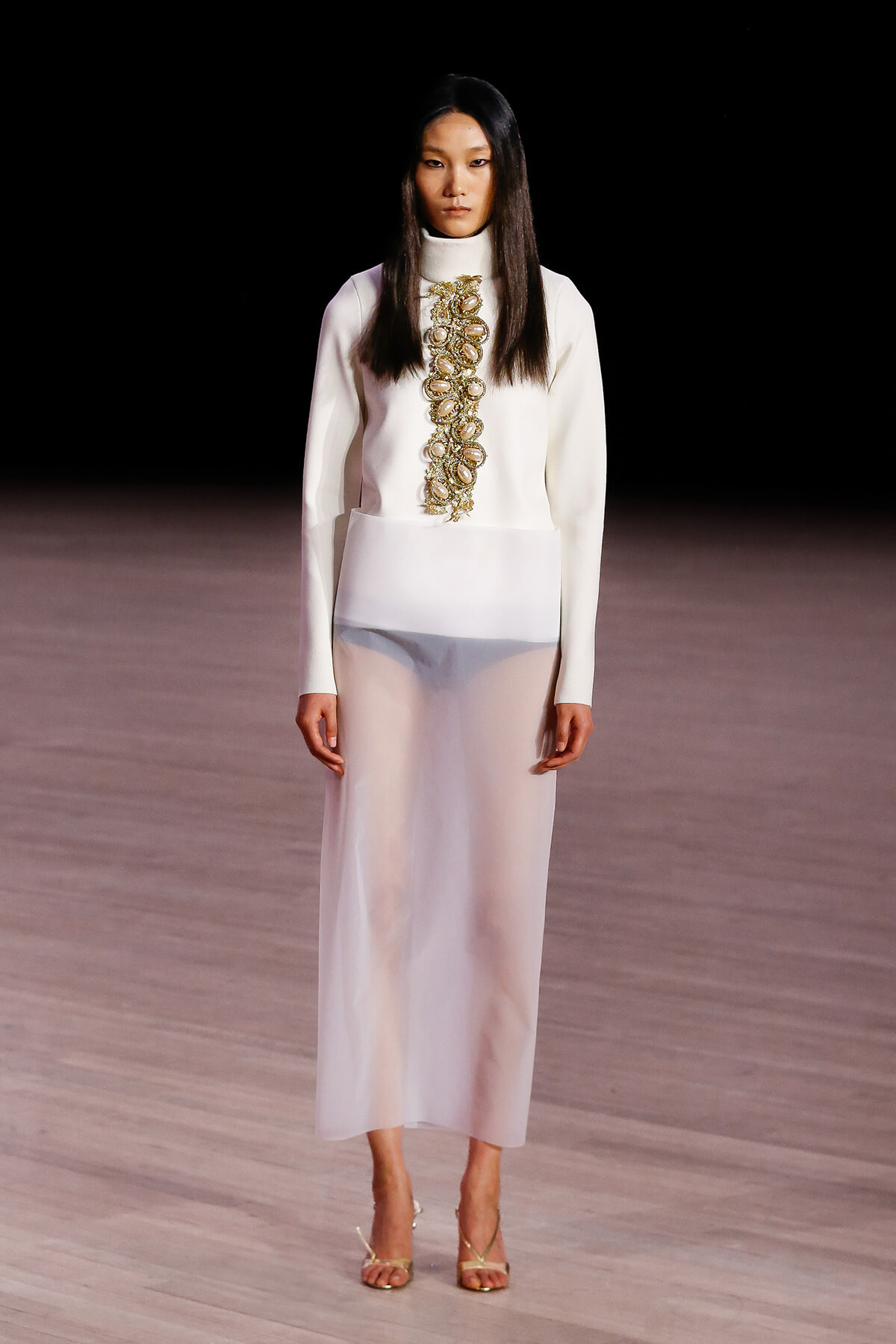 Model on a runway wearing a white high-neck top with a gold-embellished vertical appliqué and matching white skirt, gold strappy heels.