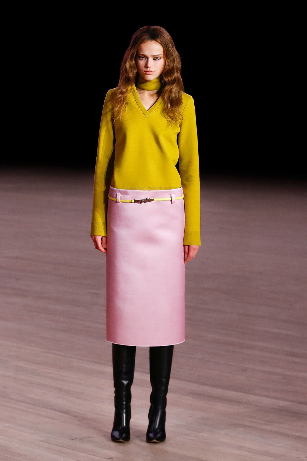 Model on a runway wearing a bright yellow V-neck sweater, pale pink midi skirt with a belt, and black knee-high boots on a wooden catwalk.