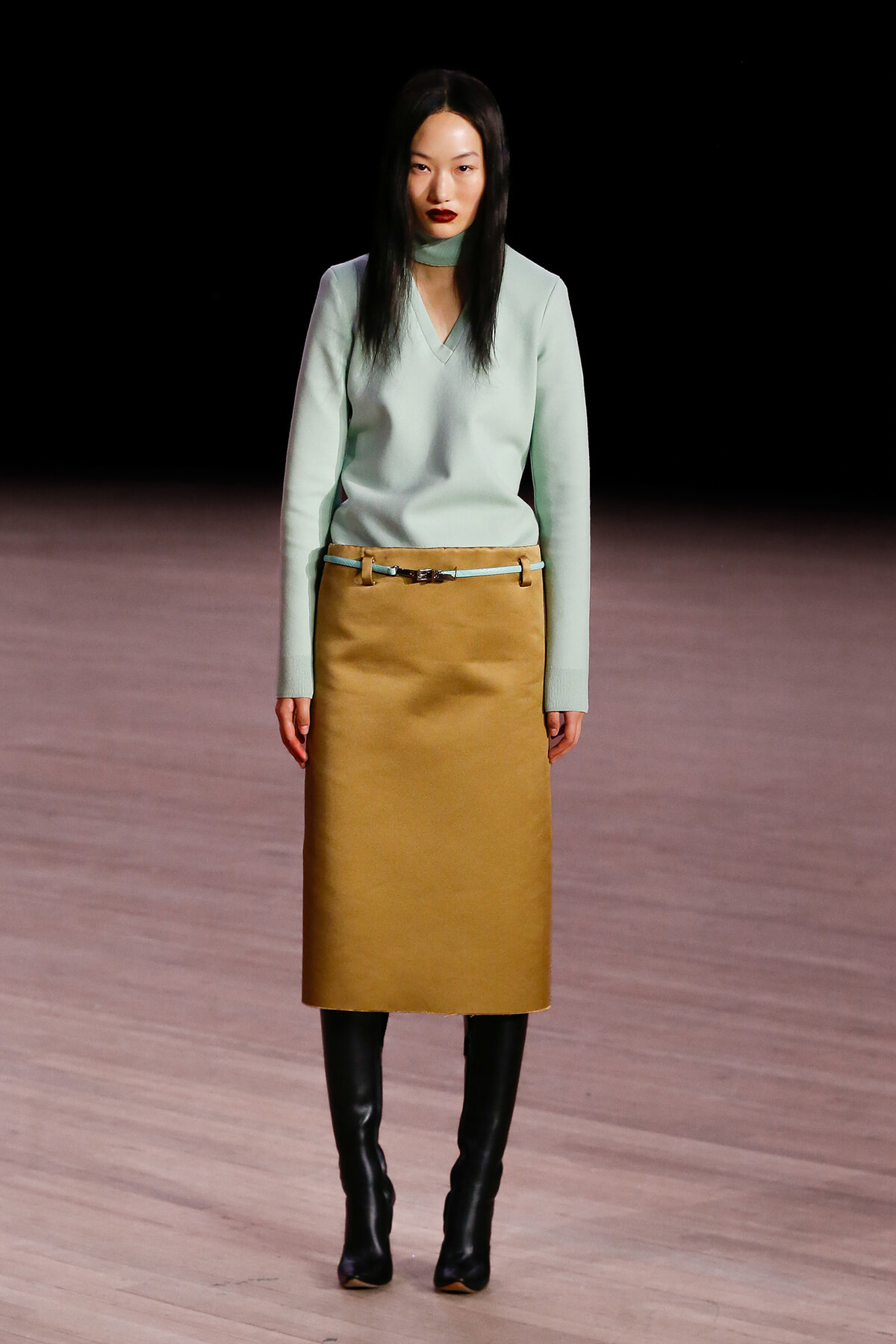 Model walking the runway in a pale mint sweater and tan midi skirt with a slim belt, paired with black knee‑high boots.
