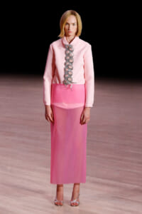 Model walking a pink monochrome outfit with a long pink skirt and pale pink blouse featuring a row of silver circular embellishments down the front, on a runway.