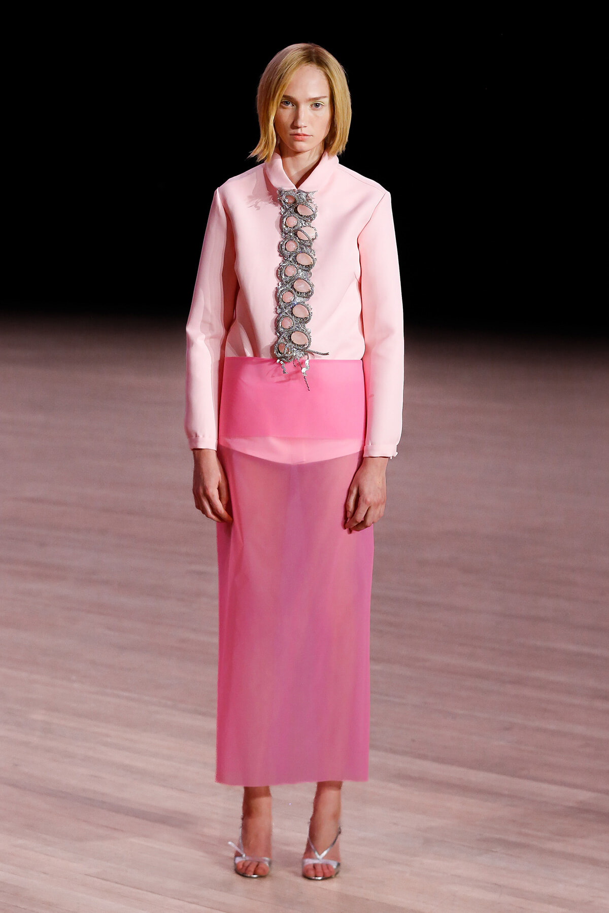 Model walking a pink monochrome outfit with a long pink skirt and pale pink blouse featuring a row of silver circular embellishments down the front, on a runway.