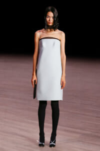 Model on a runway wearing a strapless pale blue sheath dress with brown neckline trim, black tights, and silver-heeled sandals, carrying a small black clutch.