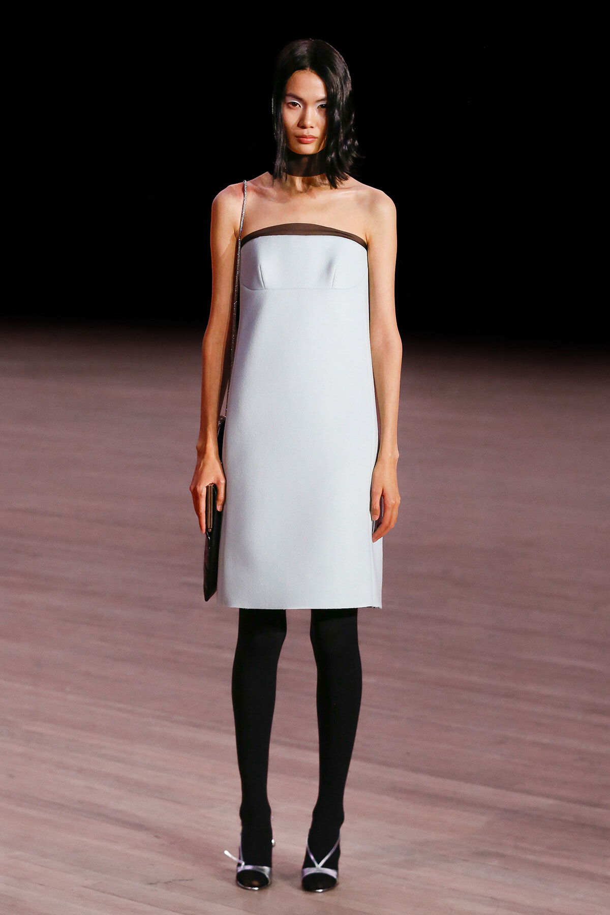 Model on a runway wearing a strapless pale blue sheath dress with brown neckline trim, black tights, and silver-heeled sandals, carrying a small black clutch.