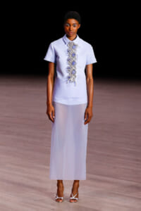 Model on a runway wearing a lavender short-sleeve blouse with an ornate beaded embroidery panel down the front and a matching sheer lavender skirt.