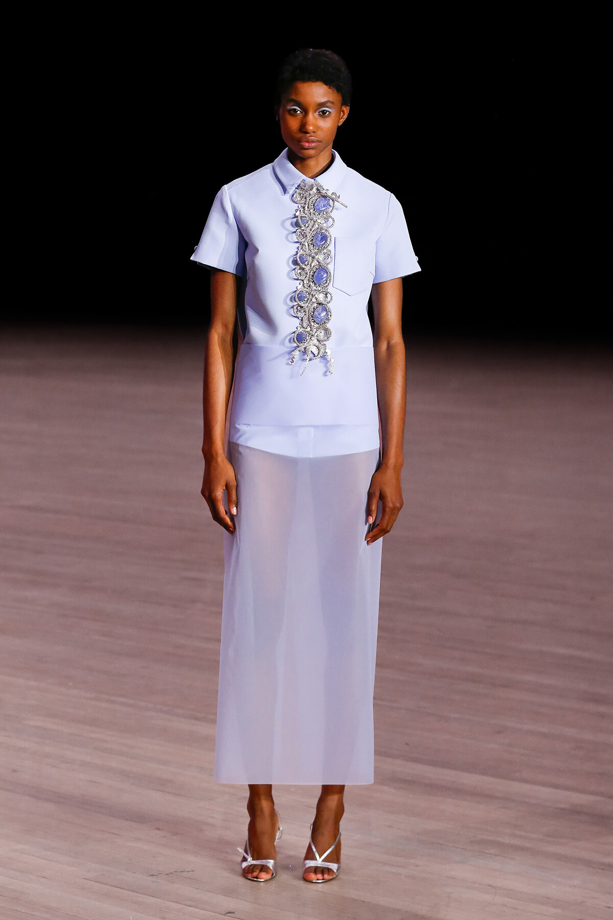 Model on a runway wearing a lavender short-sleeve blouse with an ornate beaded embroidery panel down the front and a matching sheer lavender skirt.