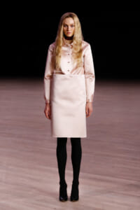 Model wearing a pale pink satin blouse tucked into a knee-length matching skirt, black tights, and black heels on a runway.