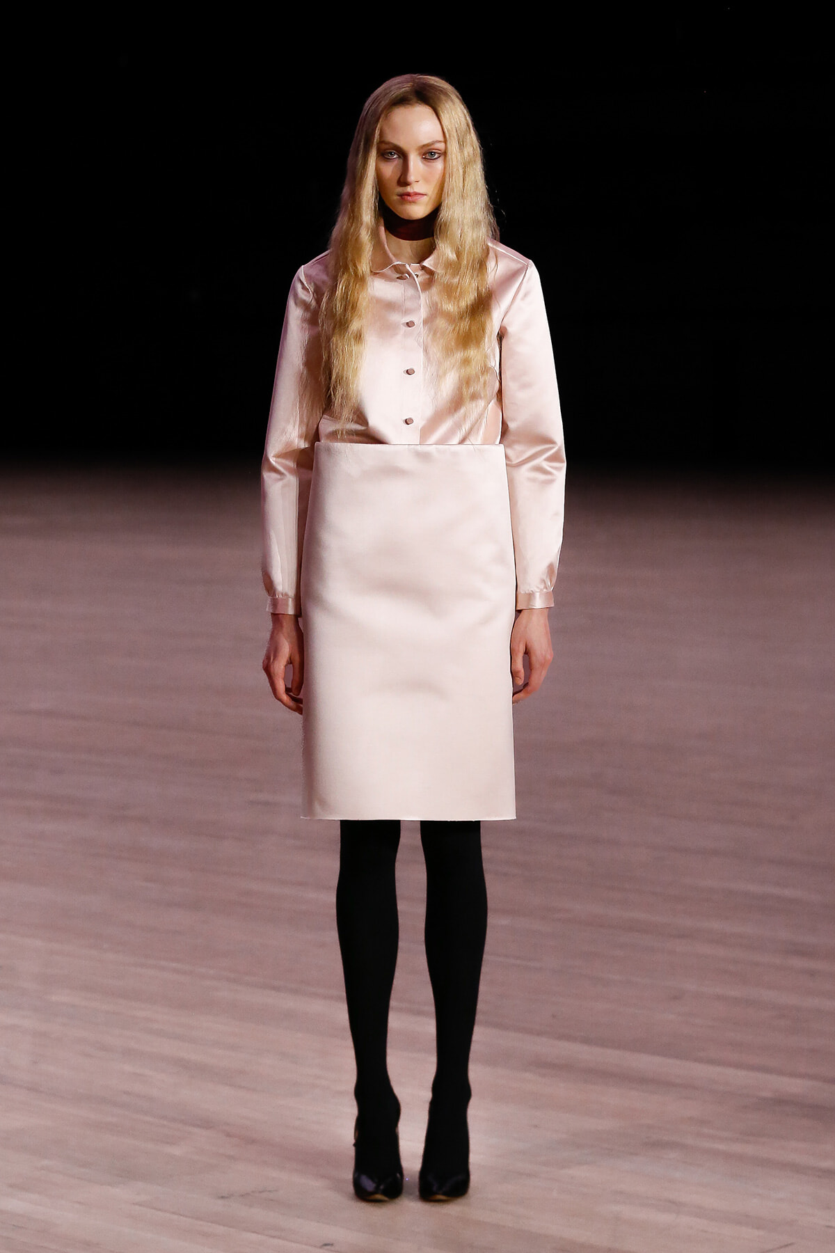 Model wearing a pale pink satin blouse tucked into a knee-length matching skirt, black tights, and black heels on a runway.