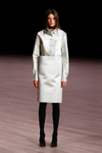 Model on a runway wearing a pale mint button-down top tucked into a knee-length skirt, with black tights and dark heels against a dark backdrop.