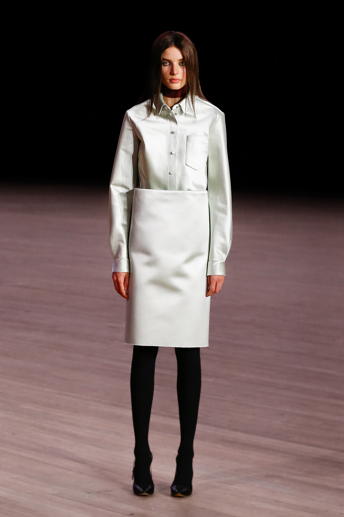 Model on a runway wearing a pale mint button-down top tucked into a knee-length skirt, with black tights and dark heels against a dark backdrop.