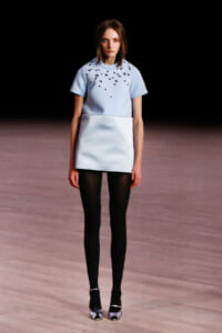 Model walking a runway in a pale blue short-sleeve top with scattered embellishments and a matching boxy mini skirt, black tights, and silver heels.