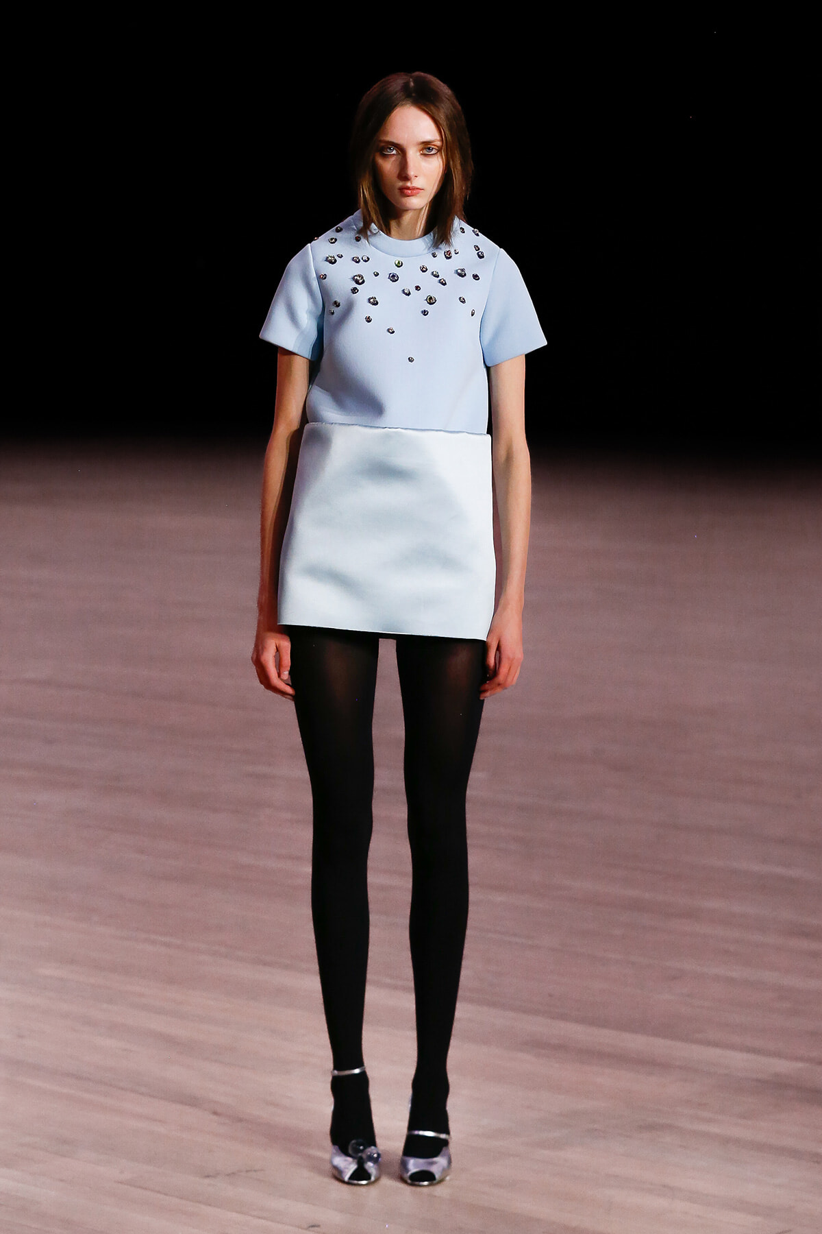Model walking a runway in a pale blue short-sleeve top with scattered embellishments and a matching boxy mini skirt, black tights, and silver heels.