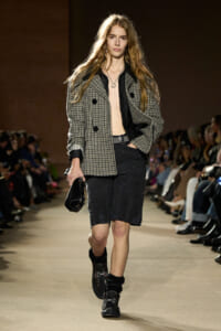 Model walking the runway in a gray plaid double-breasted coat over a black shirt, black knee-length shorts, chunky black boots, and a small black handbag; audience along the catwalk.