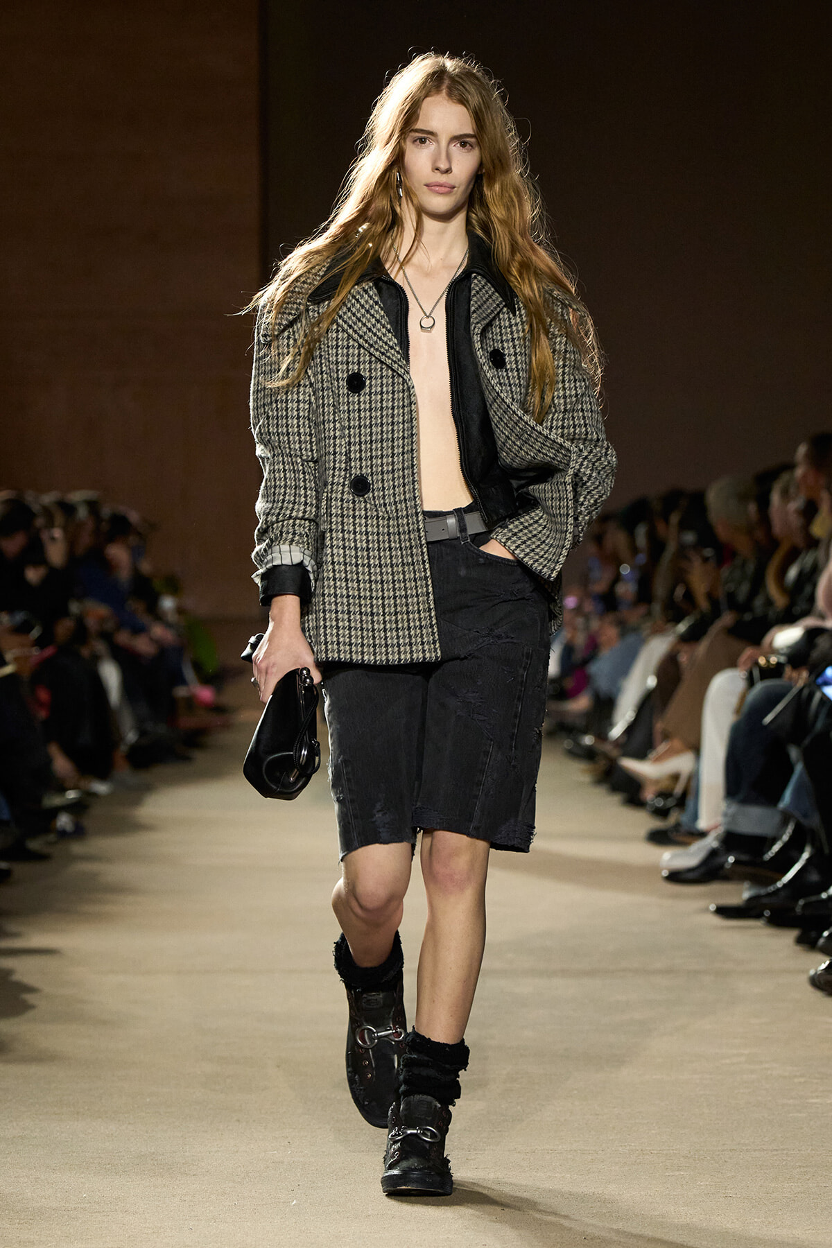 Model walking the runway in a gray plaid double-breasted coat over a black shirt, black knee-length shorts, chunky black boots, and a small black handbag; audience along the catwalk.