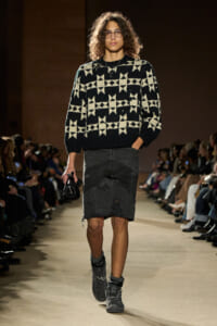 Model walks the runway in a black-and-cream geometric knit sweater with a distressed dark denim skirt and gray combat boots, carrying a small black bag.