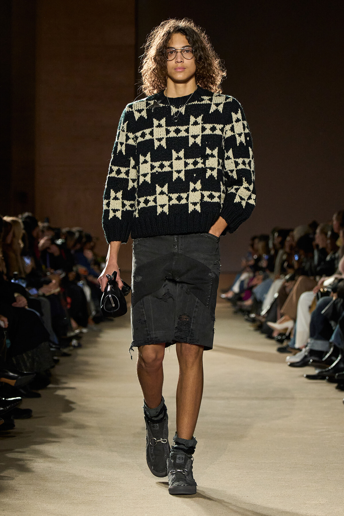 Model walks the runway in a black-and-cream geometric knit sweater with a distressed dark denim skirt and gray combat boots, carrying a small black bag.