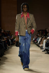 Model walking a fashion runway in a tan plaid blazer with red lapels and cuffs, pink shirt, distressed wide-leg jeans, carrying a small black bag.