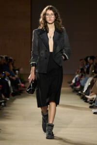 Female model walks a runway in a dark, oversized blazer, black midi skirt, and chunky gray boots, holding a small black purse and wearing glasses.
