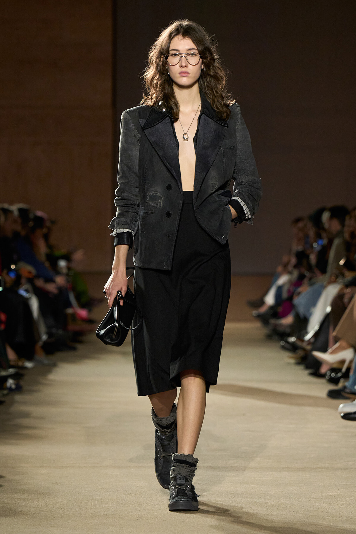 Female model walks a runway in a dark, oversized blazer, black midi skirt, and chunky gray boots, holding a small black purse and wearing glasses.