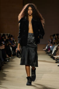 Model walking a runway in a black double-breasted coat, cropped shirtless look, and a black leather midi skirt with belt; fur-lined boots and small clutch in hand.