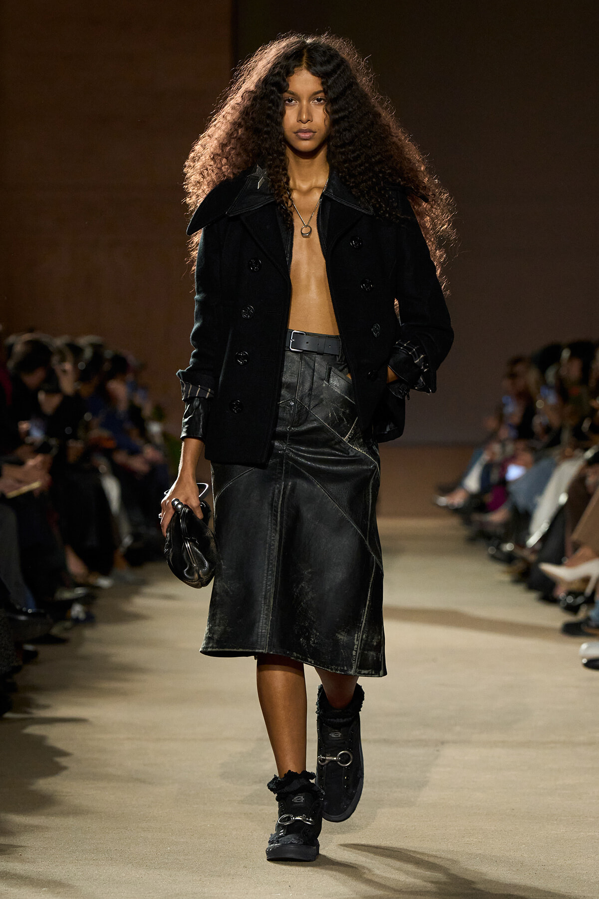 Model walking a runway in a black double-breasted coat, cropped shirtless look, and a black leather midi skirt with belt; fur-lined boots and small clutch in hand.