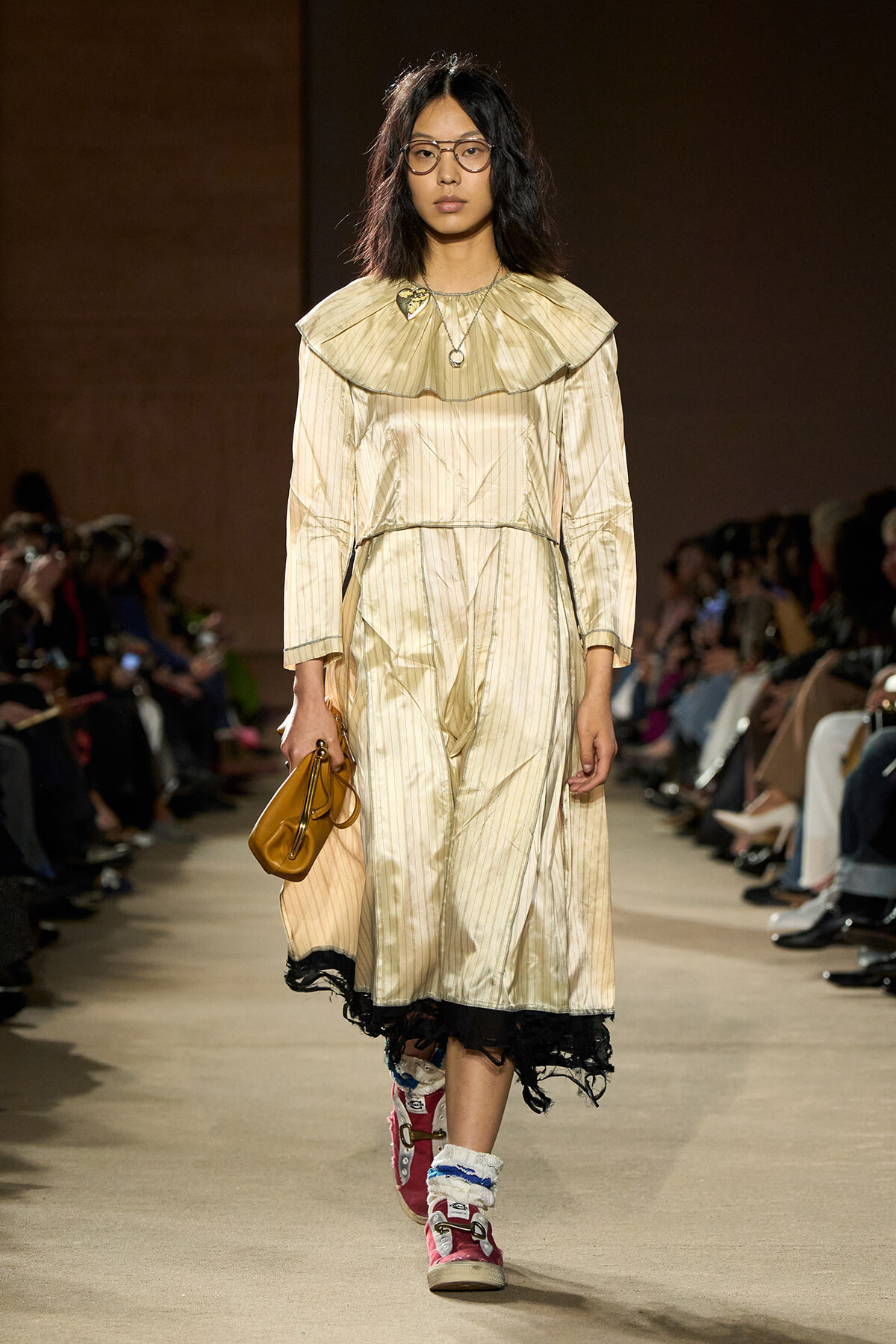 Model on runway wearing a cream striped dress with oversized ruffled collar, black lace trim, and a brown handbag, audience in background.
