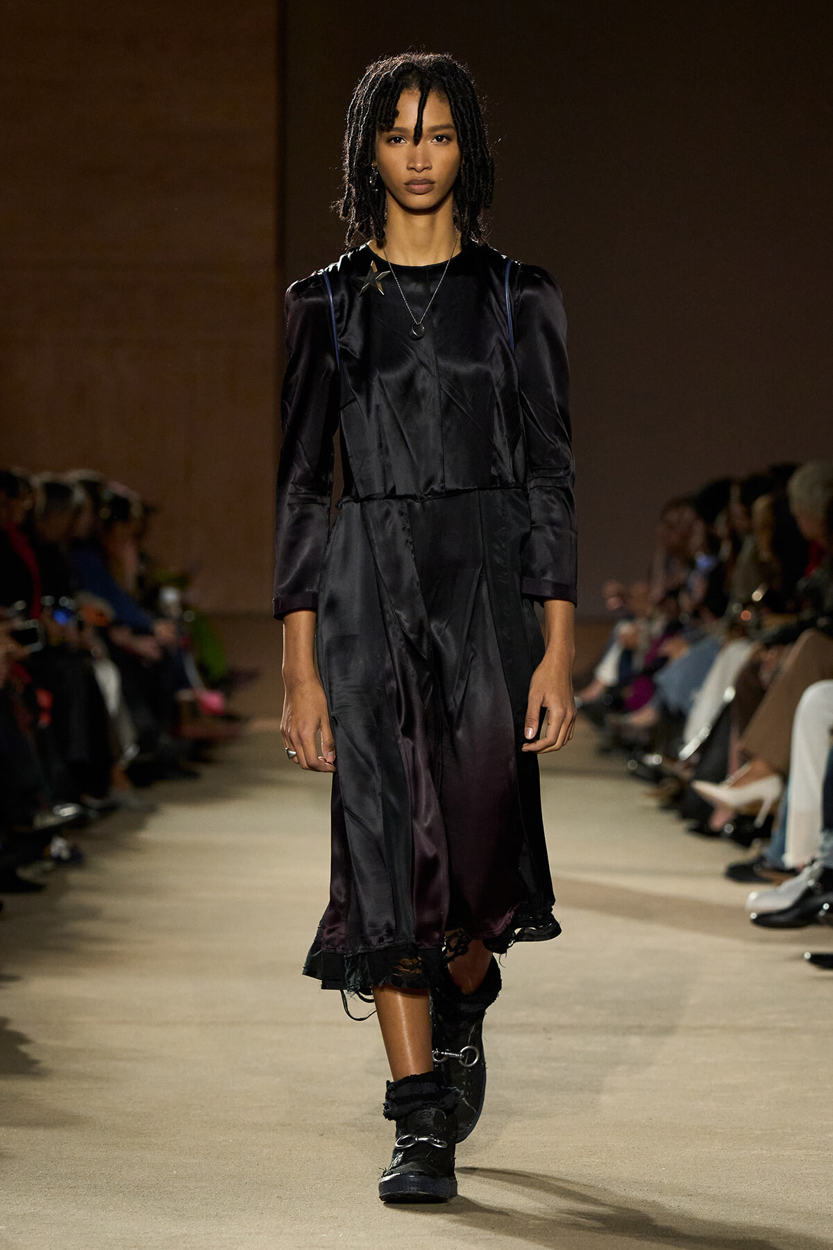 Model walking a runway in a long black satin dress with puffed shoulders and black boots, audience on both sides.