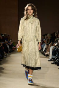 Model walking a runway in a long beige striped dress with a high collar, holding a small mustard handbag.