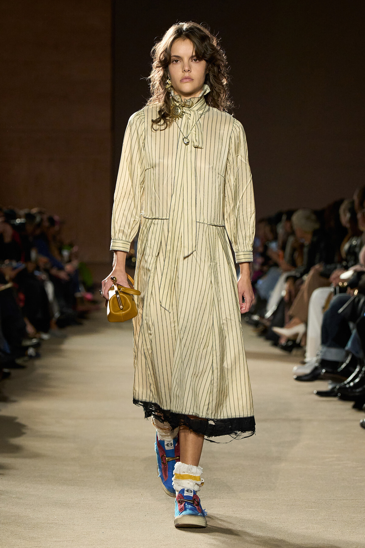 Model walking a runway in a long beige striped dress with a high collar, holding a small mustard handbag.