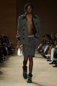 Male model walks the runway in a gray plaid oversized coat, open at the chest, paired with distressed gray shorts and dark boots with a white clutch edge accessory.